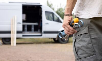 A hand of a man is holding several tools. The background shows a white van with an open side door and wooden slats leaning against it.